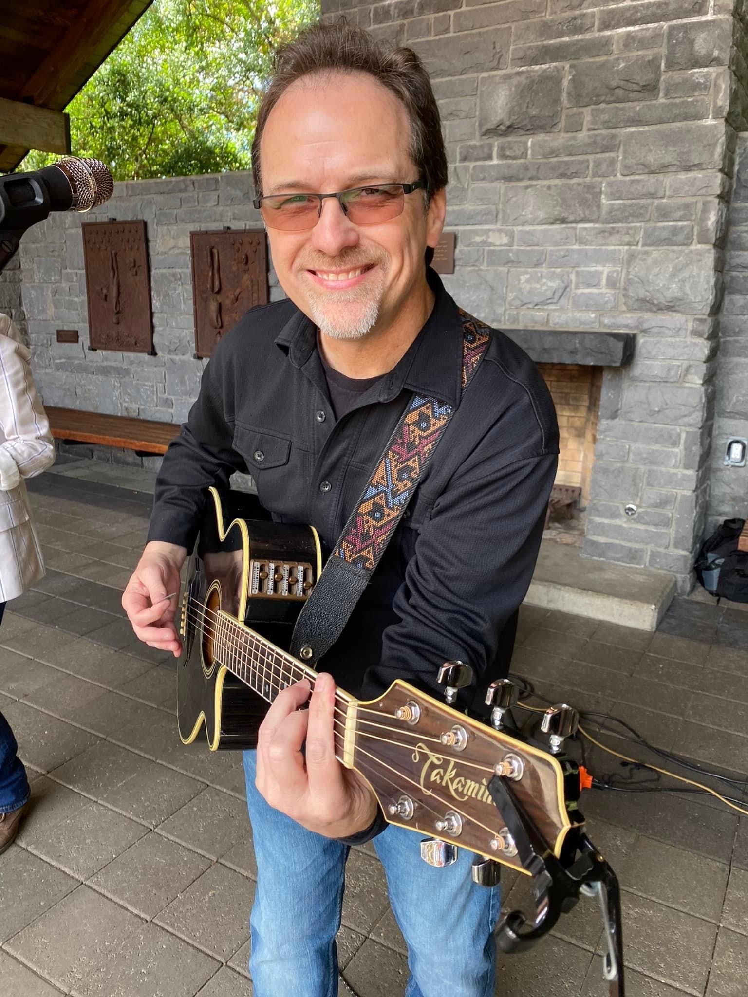 A man is playing an acoustic guitar in front of a stone wall.