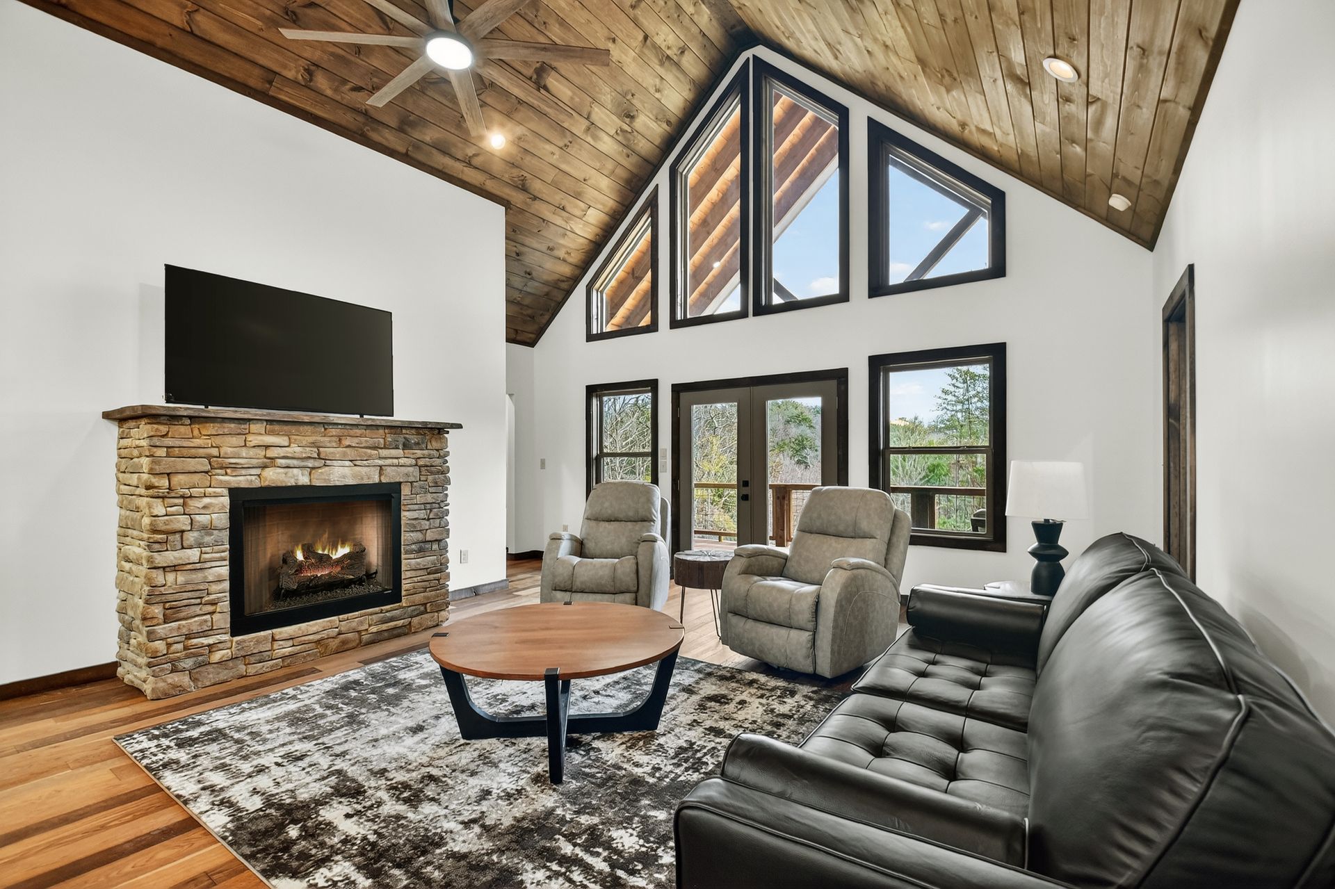 Modern living room with vaulted wood ceiling, stone fireplace, black sofa, and large windows