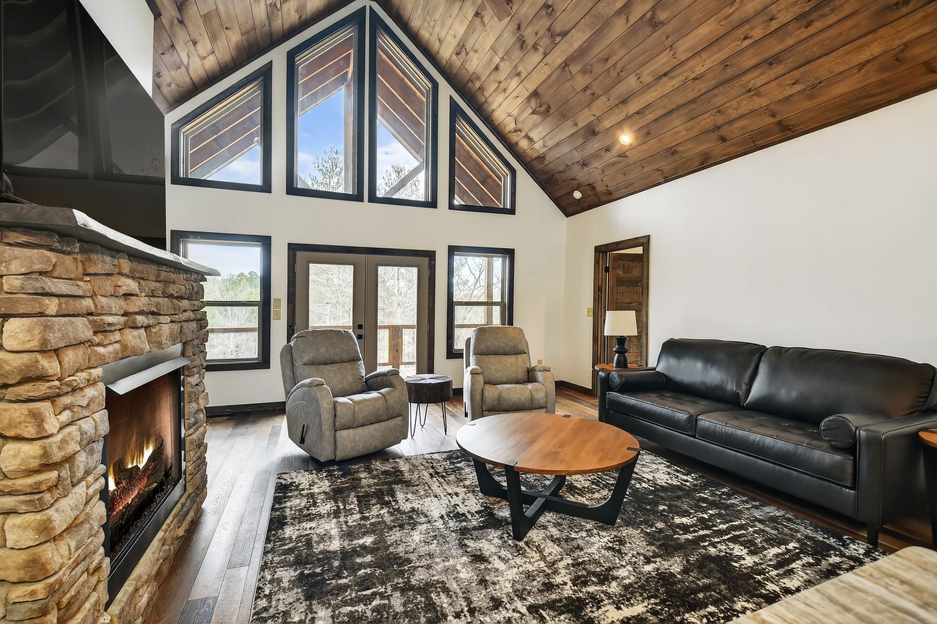 Spacious living room with stone fireplace, vaulted wood ceiling, black sofa, and two armchairs by tall windows.