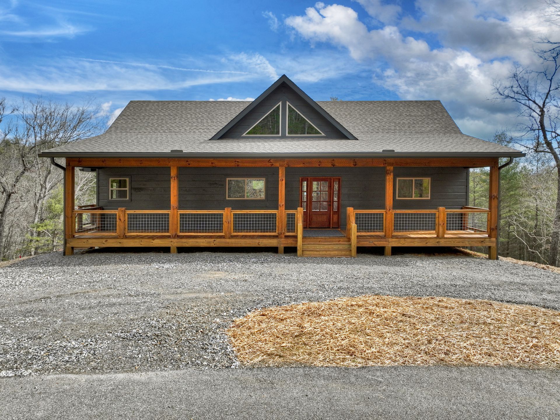 Wooden cabin with a front porch and stone chimney, set on a gravel lot under a partly cloudy blue sky