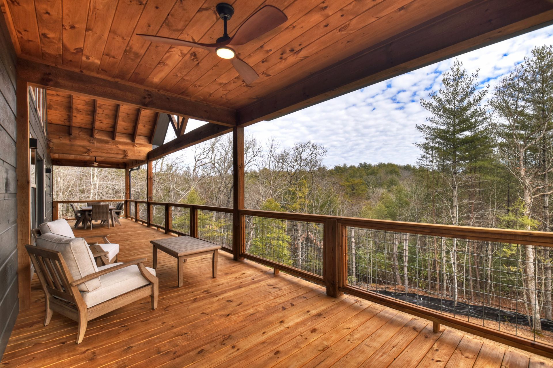 Covered wooden porch with chairs, railing, and forest view under a rustic ceiling fan