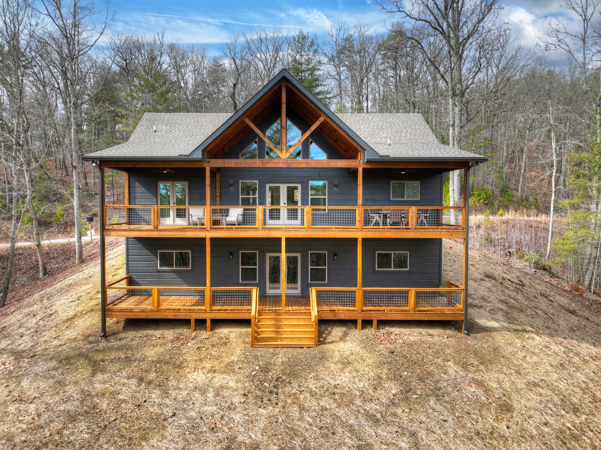Two-story rustic cabin with wraparound porch in a wooded clearing under a blue sky