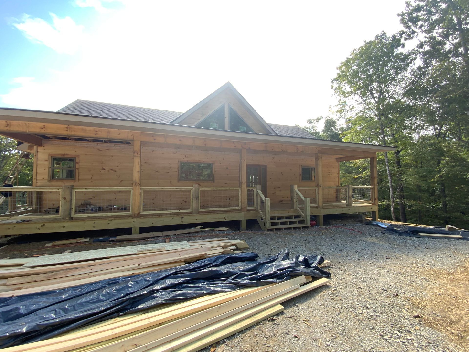 Log cabin-style house under construction with porch, gravel driveway, and lumber stacked in front