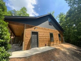 A wooden house with a black roof is sitting on top of a dirt hill surrounded by trees.