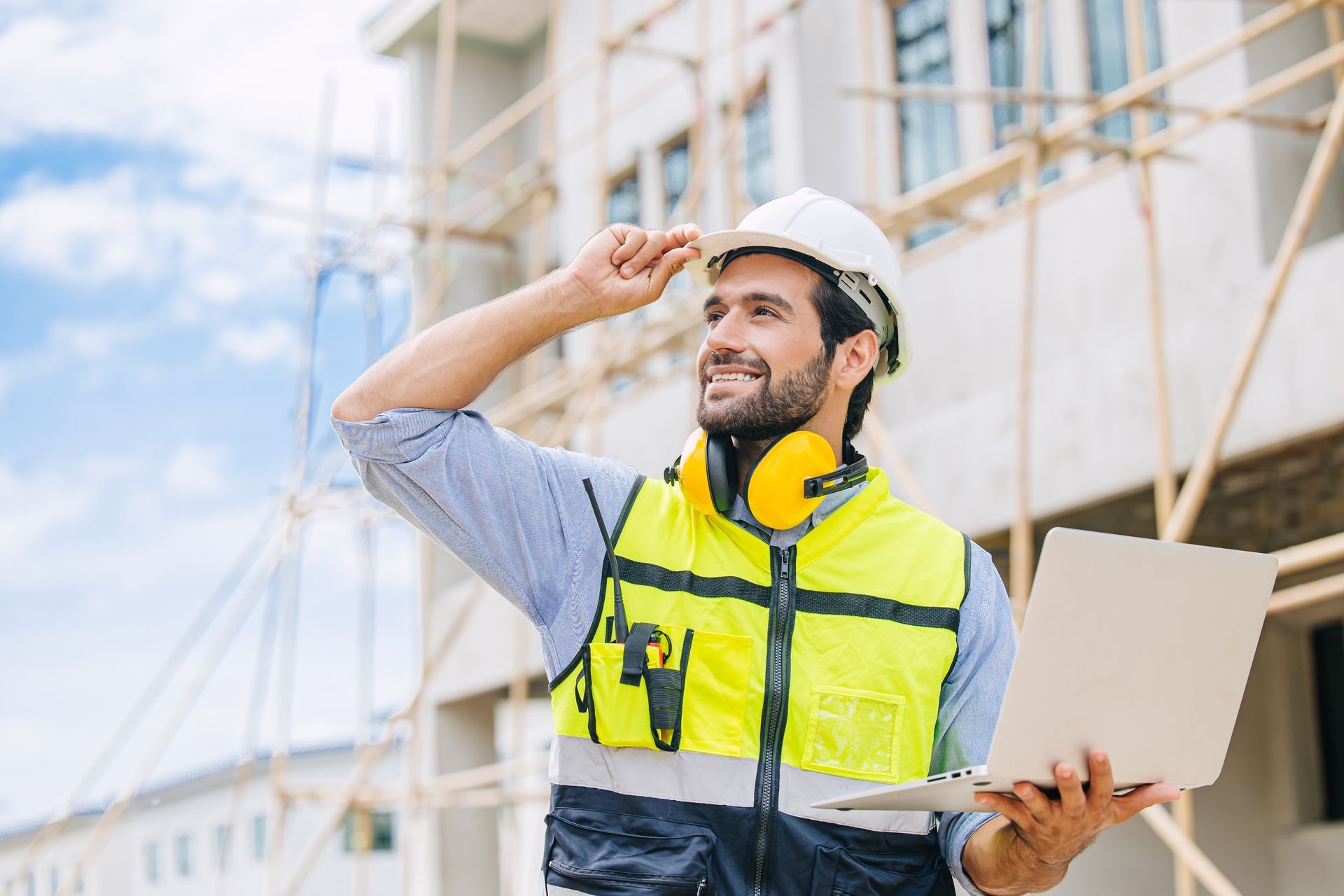 A construction worker is holding a laptop computer in front of a building under construction.