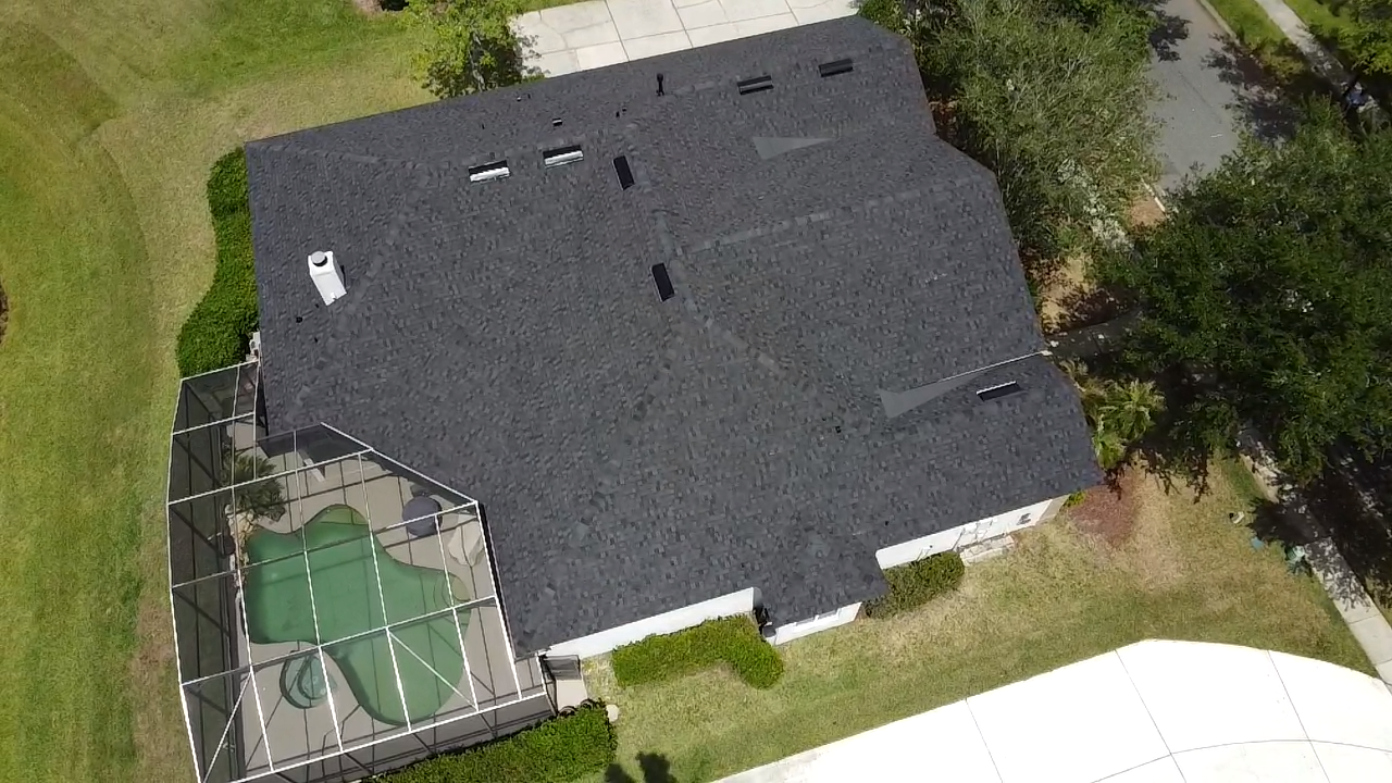 Aerial view of a house with a dark roof, backyard patio, and a fenced green tennis court.