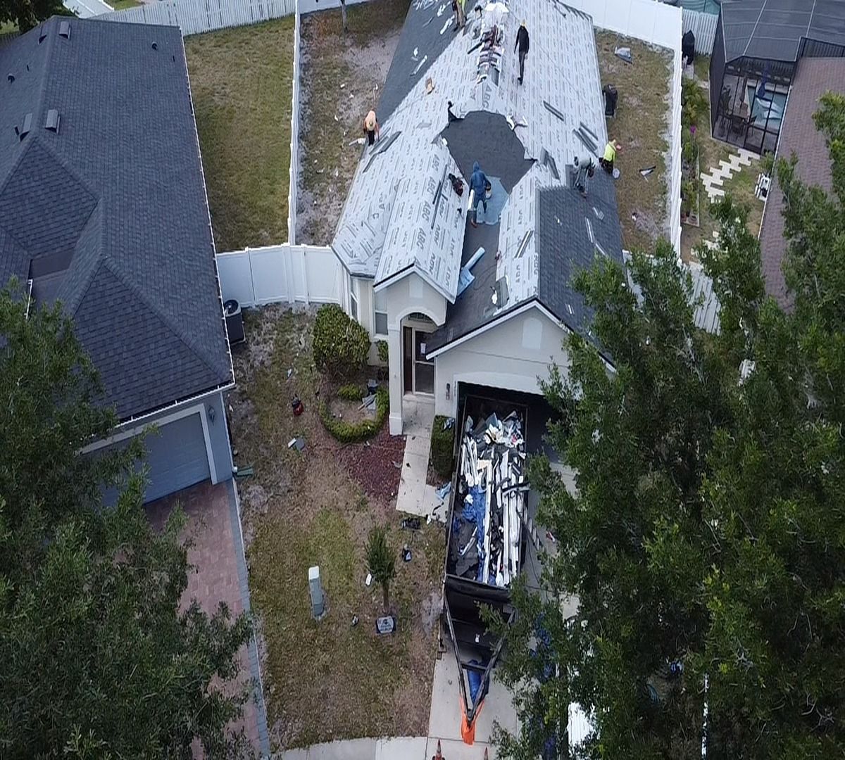 Aerial view of a damaged house roof with a large hole, debris, and nearby trees and neighboring homes.