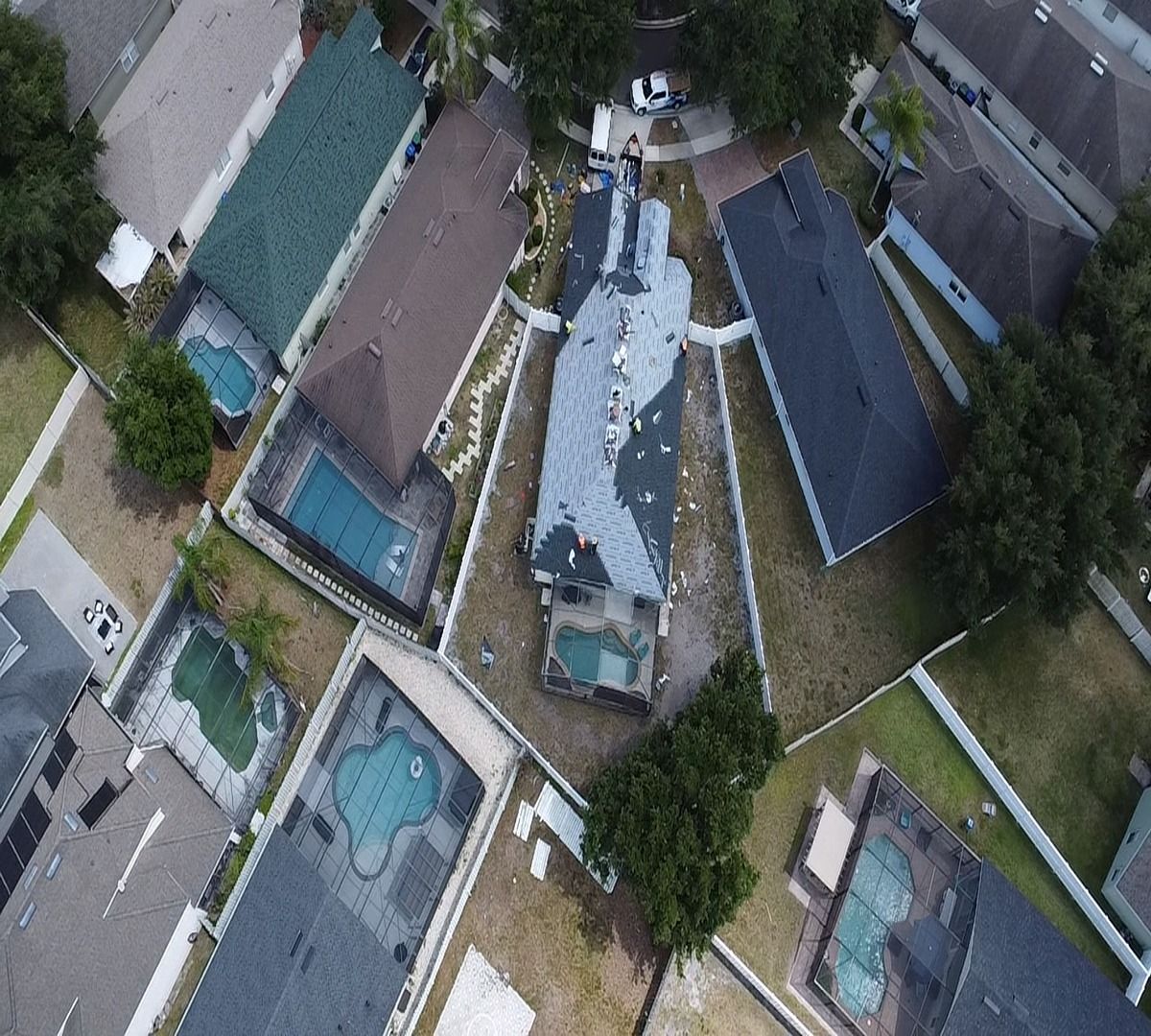 Aerial view of a house with a blue roof, swimming pools, and surrounding trees on a suburban lot