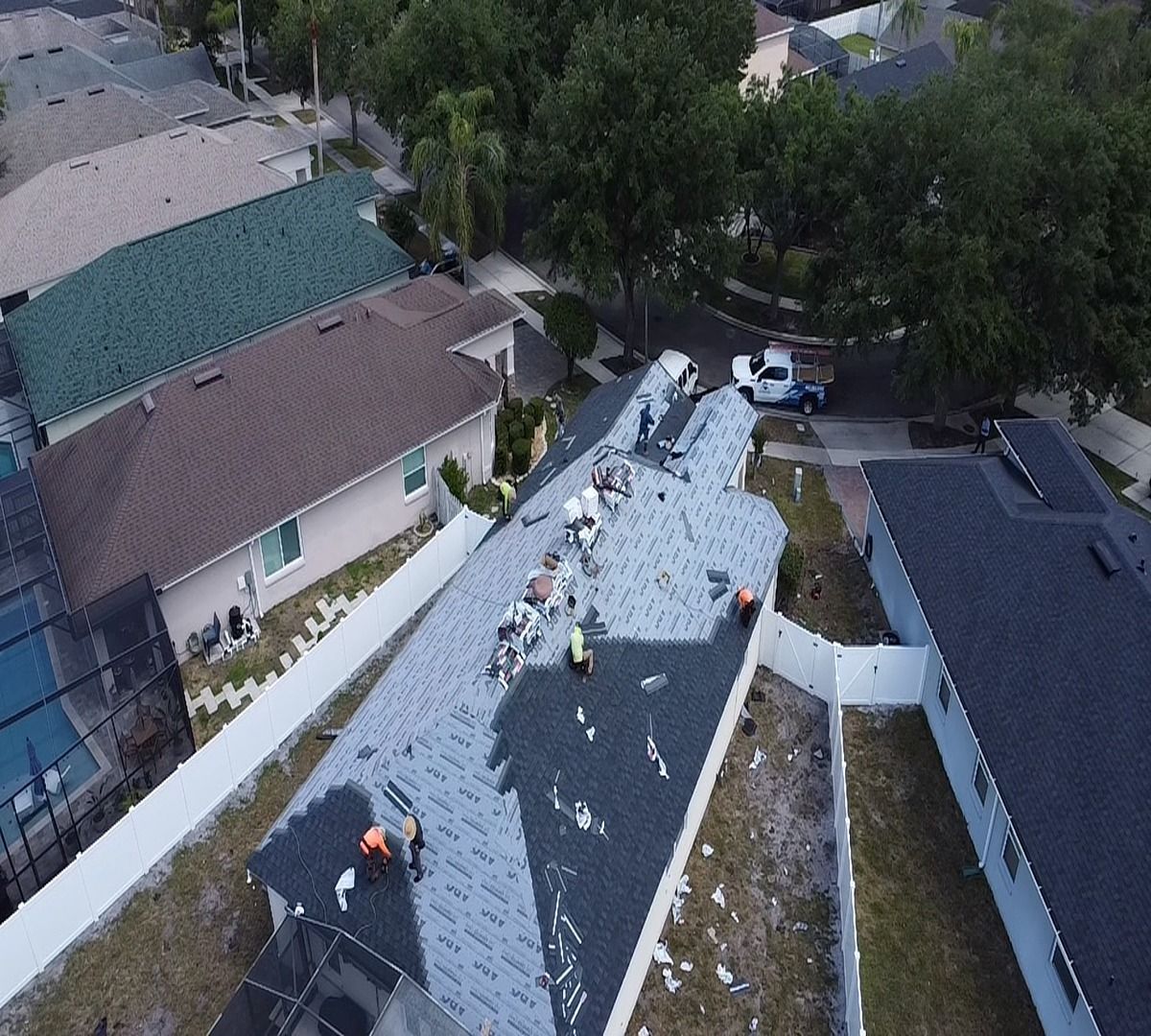 Workers on a shingled roof beside trees and houses, viewed from above.