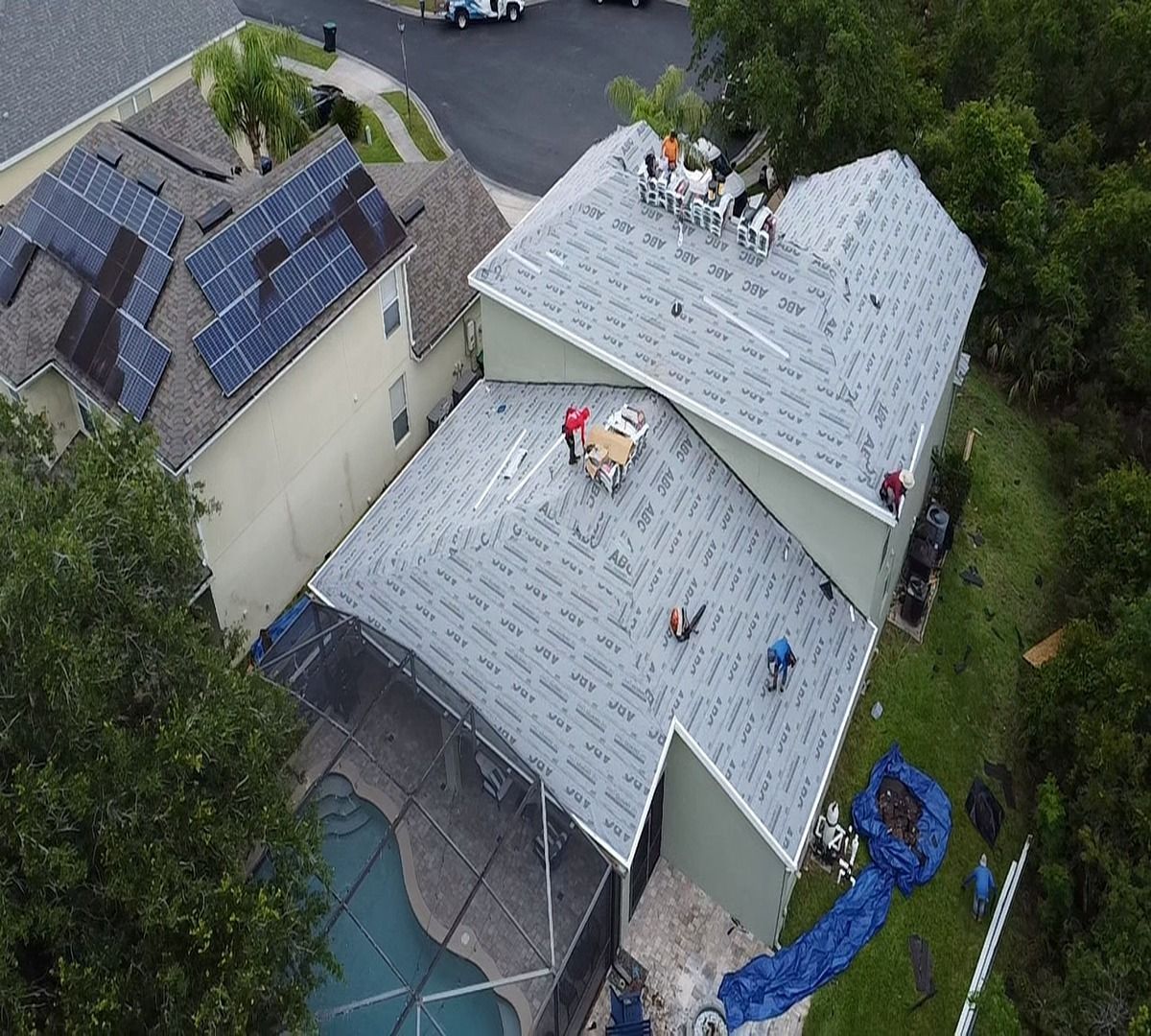 Aerial view of workers replacing a light gray roof on a house beside another with solar panels.