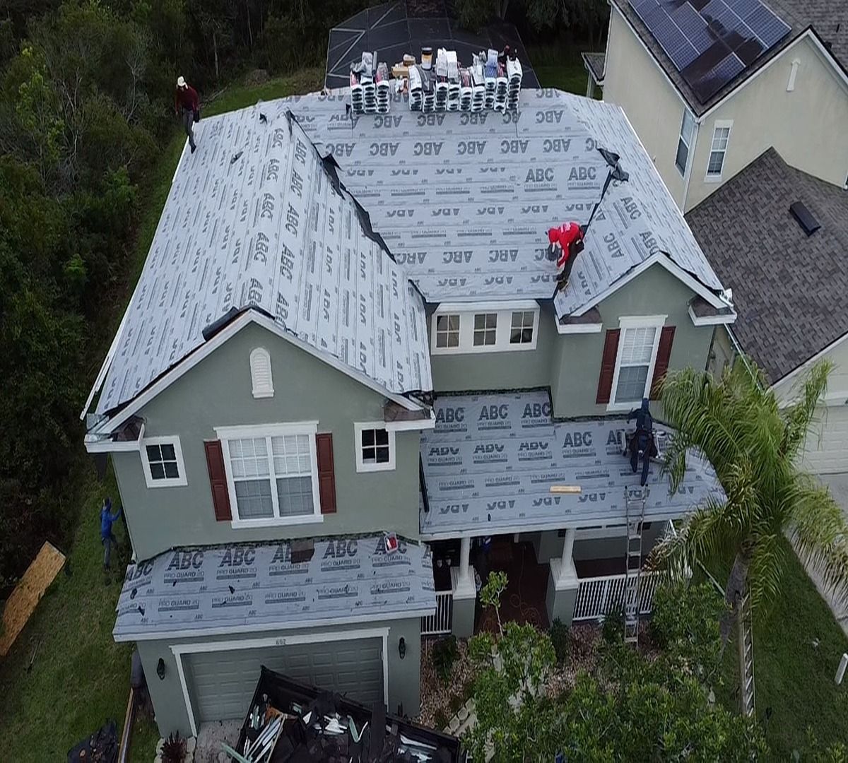 Aerial view of a house under roof repair, with white underlayment and workers on the roof.