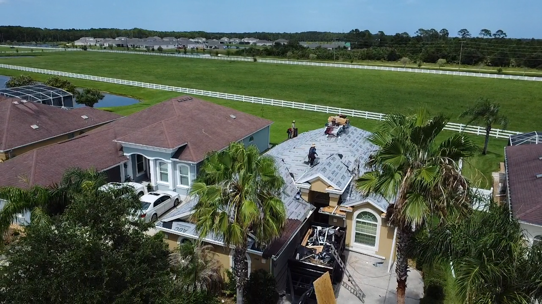 Aerial view of a suburban home with red roof, palm trees, and a wide green field beyond the backyard.
