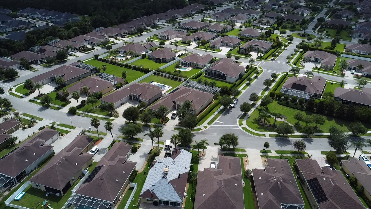 Aerial view of a suburban neighborhood with rows of houses, green lawns, and tree-lined streets.