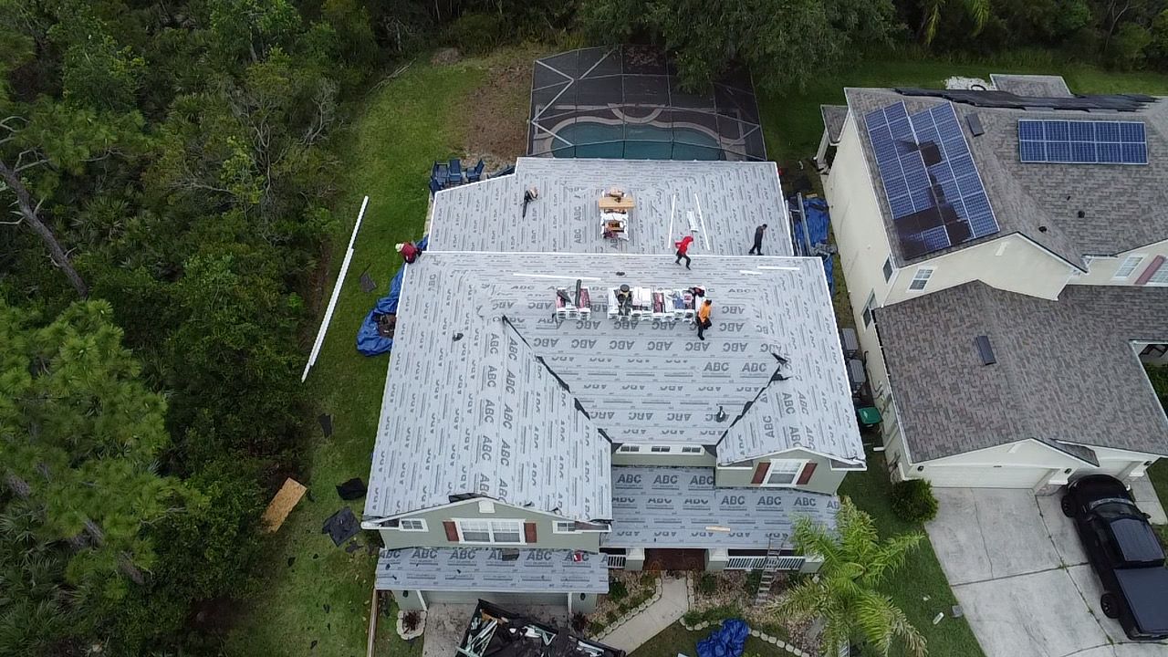 Aerial view of a white roof with workers near skylights, beside trees and neighboring houses.