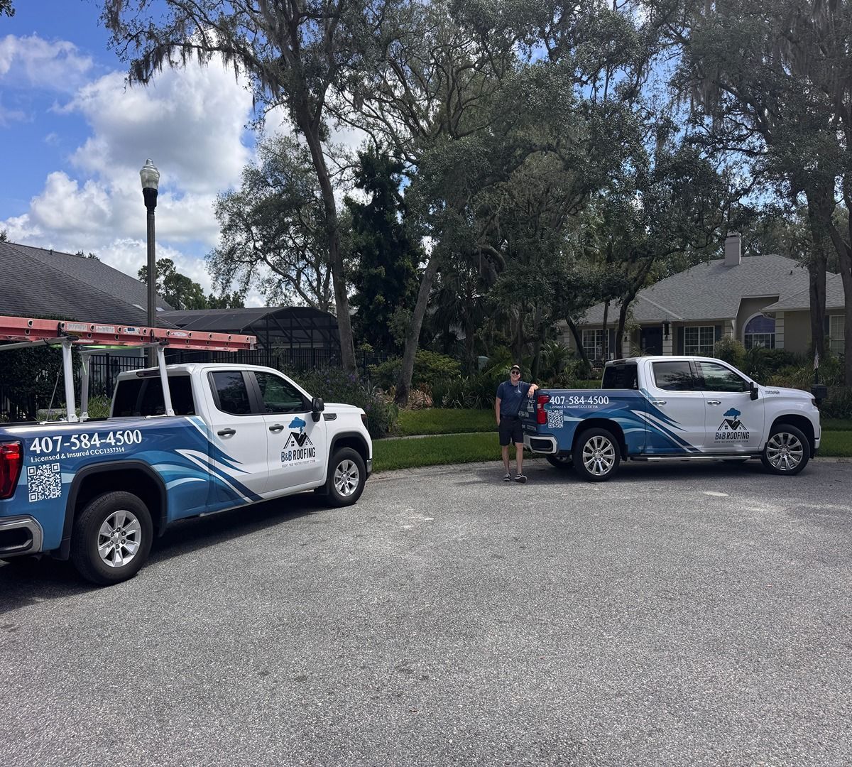 Two blue-and-white utility trucks parked on a gravel lot near houses and trees.