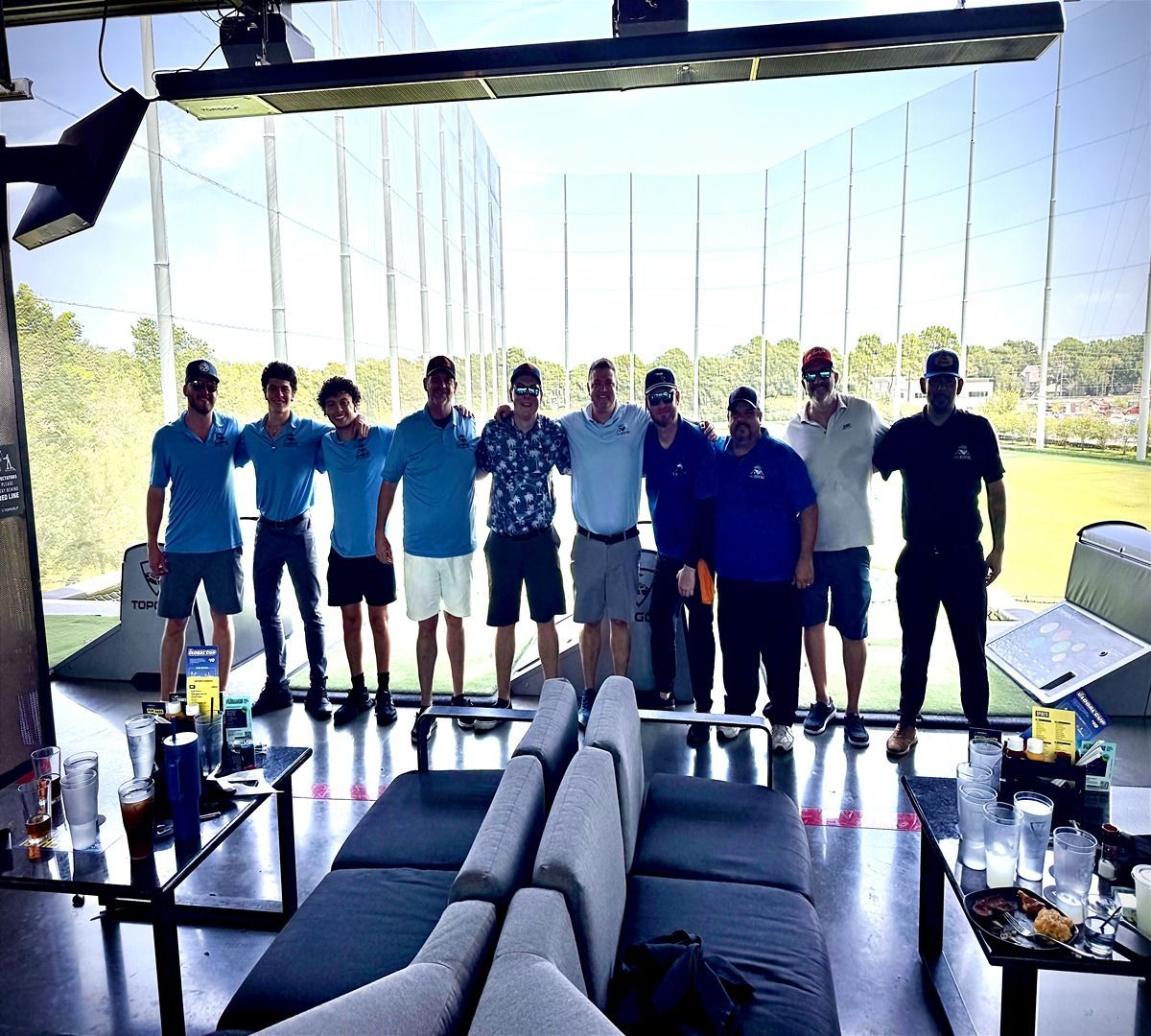 Group of people standing on an outdoor patio with lake view, behind dark lounge seating and tables.