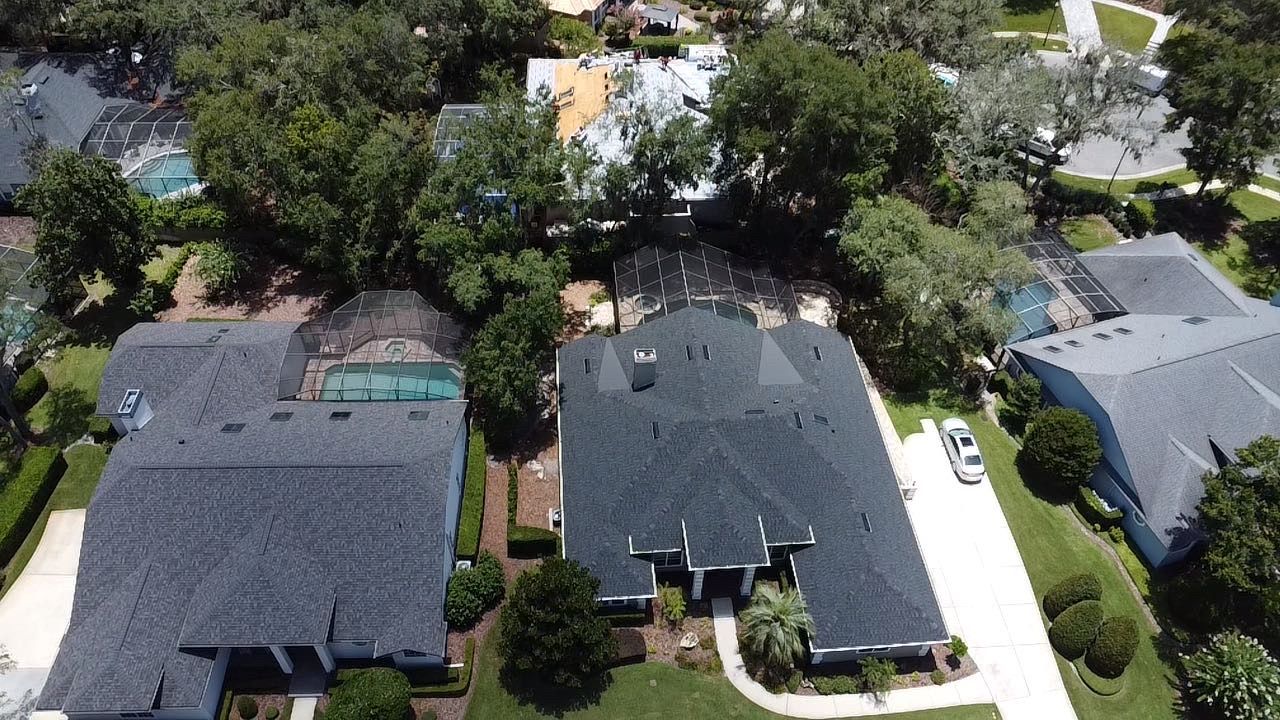 Aerial view of suburban homes with dark roofs, trees, driveways, and a swimming pool.