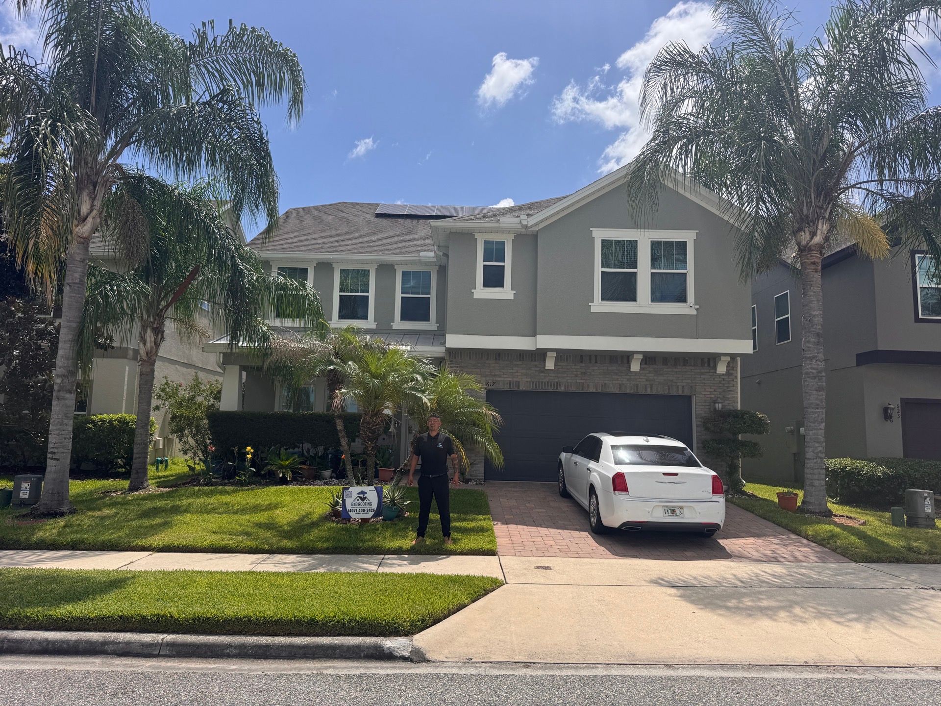Two-story suburban house with two people standing in the front yard and a white car in the driveway