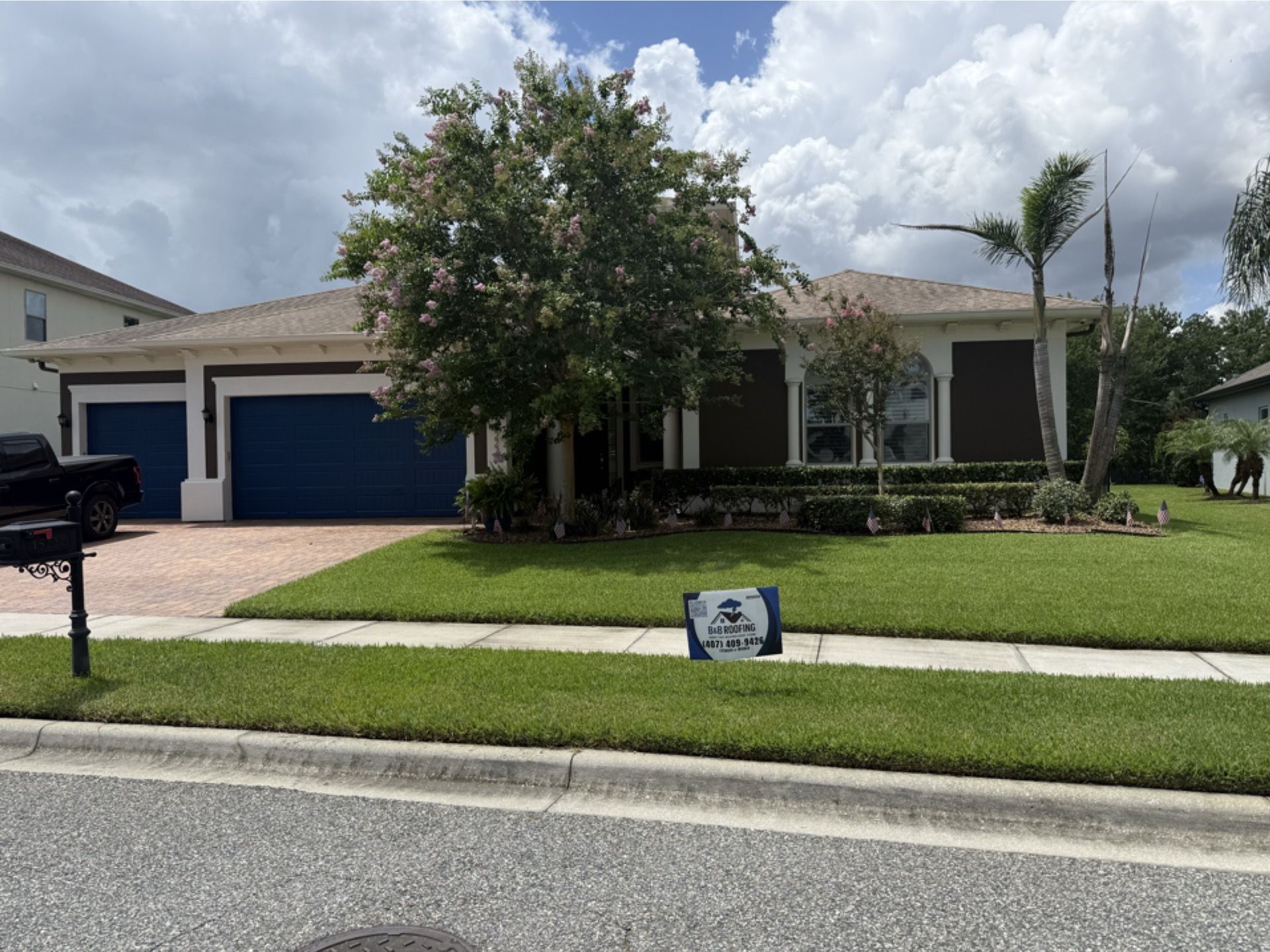 Suburban single-story house with blue garage door, palm trees, and a lawn sign by the sidewalk.