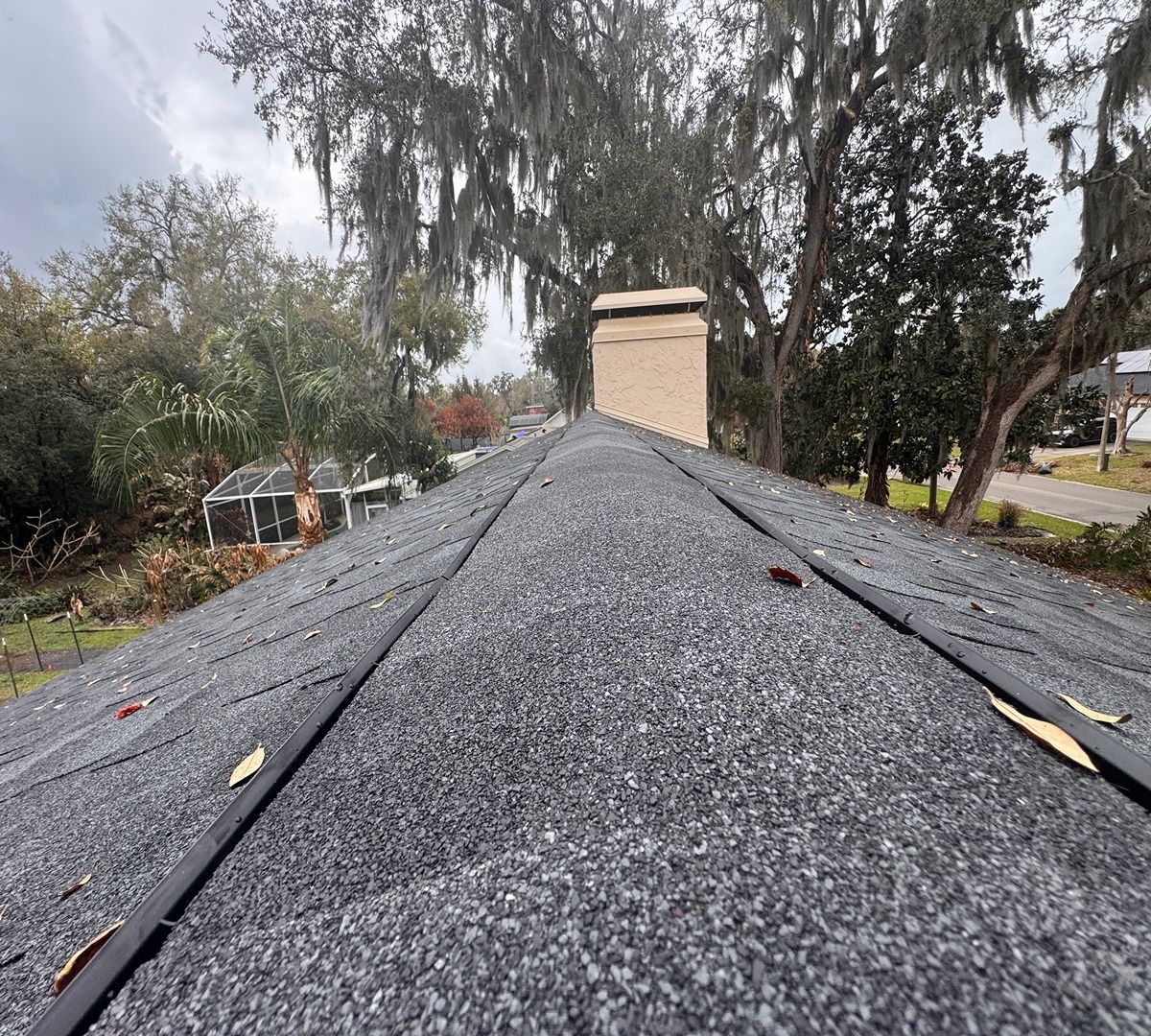 Peaked gray shingle roof viewed from the ridge, with trees and a chimney in the background
