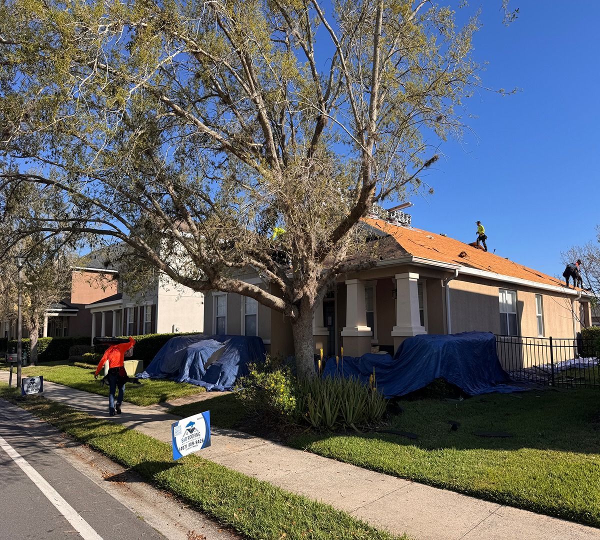 Single-story house with orange roof, covered by blue tarps, while a worker mows the front lawn.