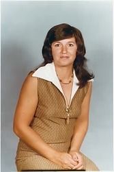 Woman seated against a gray backdrop, wearing a tan sleeveless dress with a white collar.