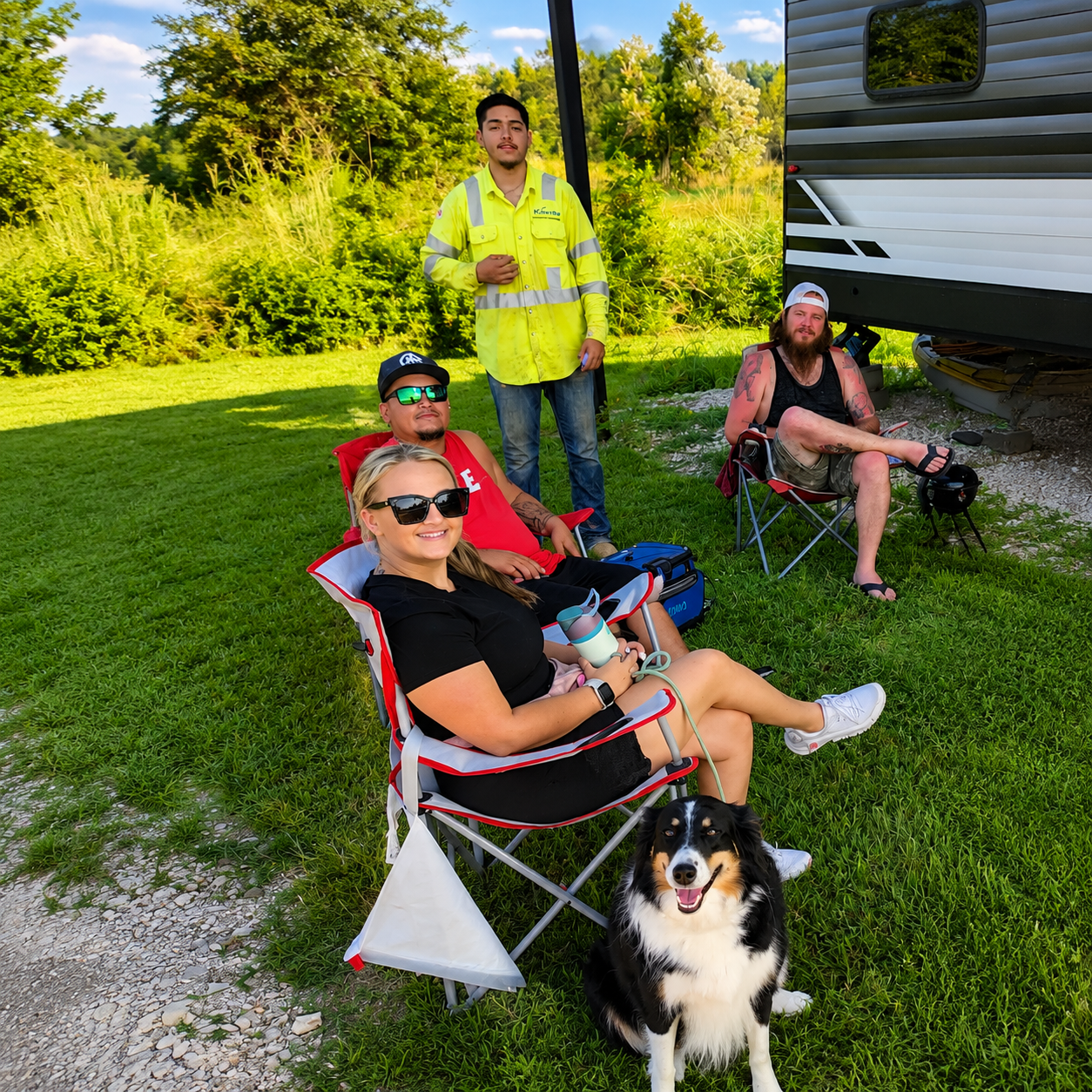 People relaxing on lawn chairs with a dog beside an RV in a sunny grassy campsite