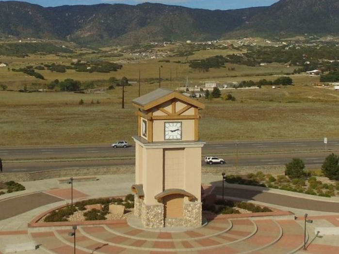 Clock tower monument in a plaza beside a road, with mountains and open fields in the background
