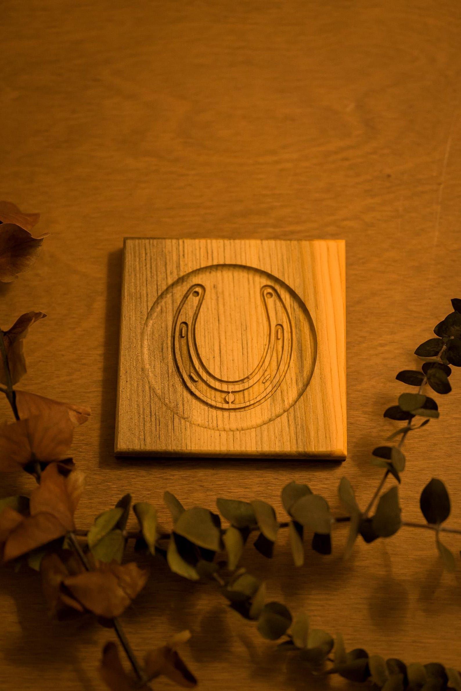 Wooden tile with a circular carved symbol, surrounded by dried eucalyptus leaves on a warm wooden surface.