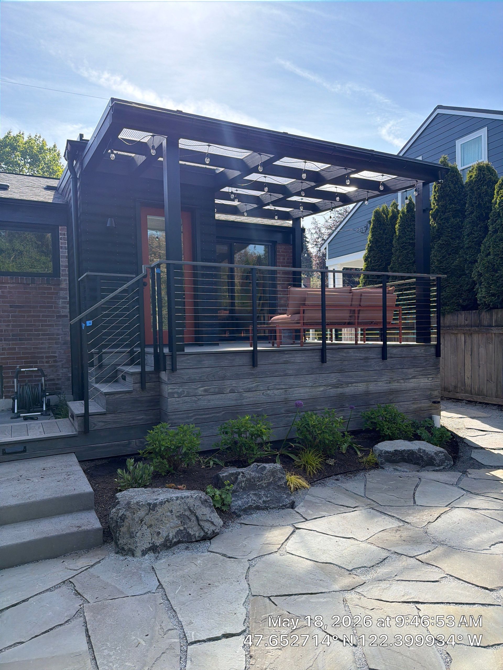 Wooden backyard deck with dining table, cushioned bench, chairs, and lush greenery beside a fence