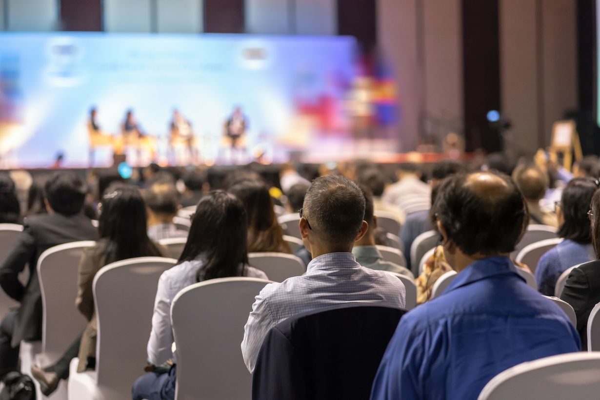 Audience seated at a conference, facing a blurred stage with speakers under colorful lights