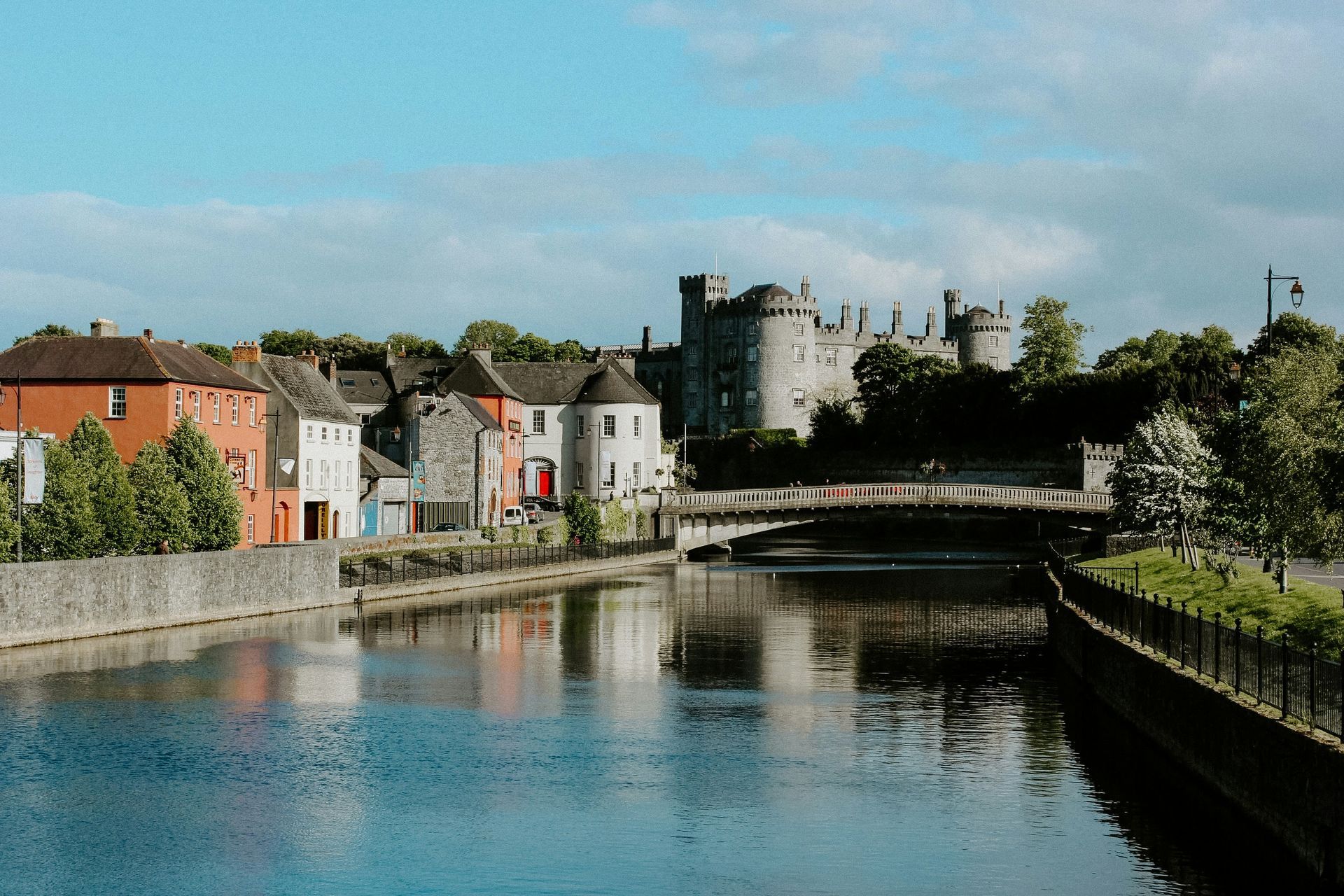 Riverfront town with stone castle, colorful houses, and calm water under a blue sky