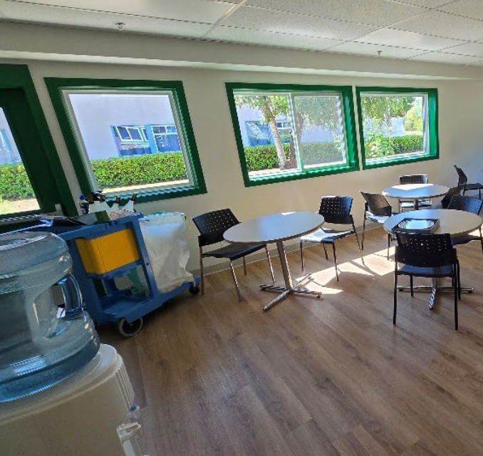 Lunchroom with tables, chairs, and large green-framed windows. A water cooler and cleaning cart are also present.
