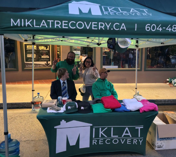 People at a Miklat Recovery booth under a green tent. Website and phone number visible.