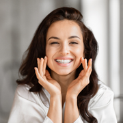 Smiling woman in a white shirt touching her cheeks, looking at the camera indoors