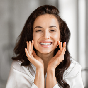 Smiling woman in a white shirt touching her cheeks, looking at the camera indoors