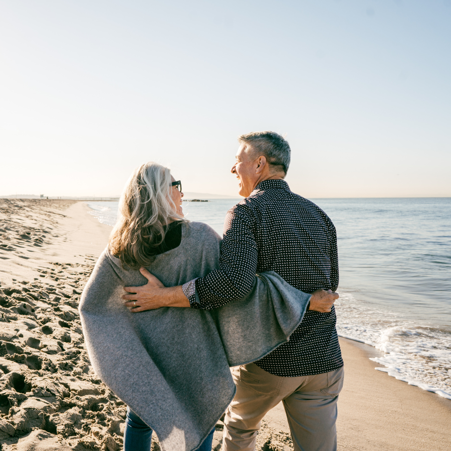 Couple walking arm-in-arm on a sandy beach at sunset, smiling toward the ocean.