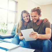 Two people smiling on a couch, looking at a tablet in a bright living room