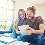 Two people smiling on a couch, looking at a tablet in a bright living room