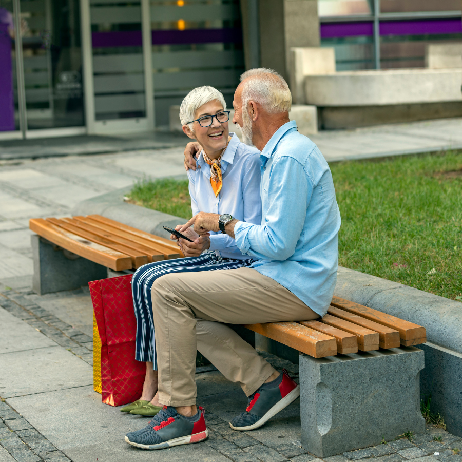 Two older adults chatting on a wooden bench outside a building, with a red bag beside them.