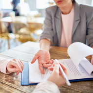 Colleagues reviewing paperwork at a wooden table, with one person gesturing and another holding a pen.