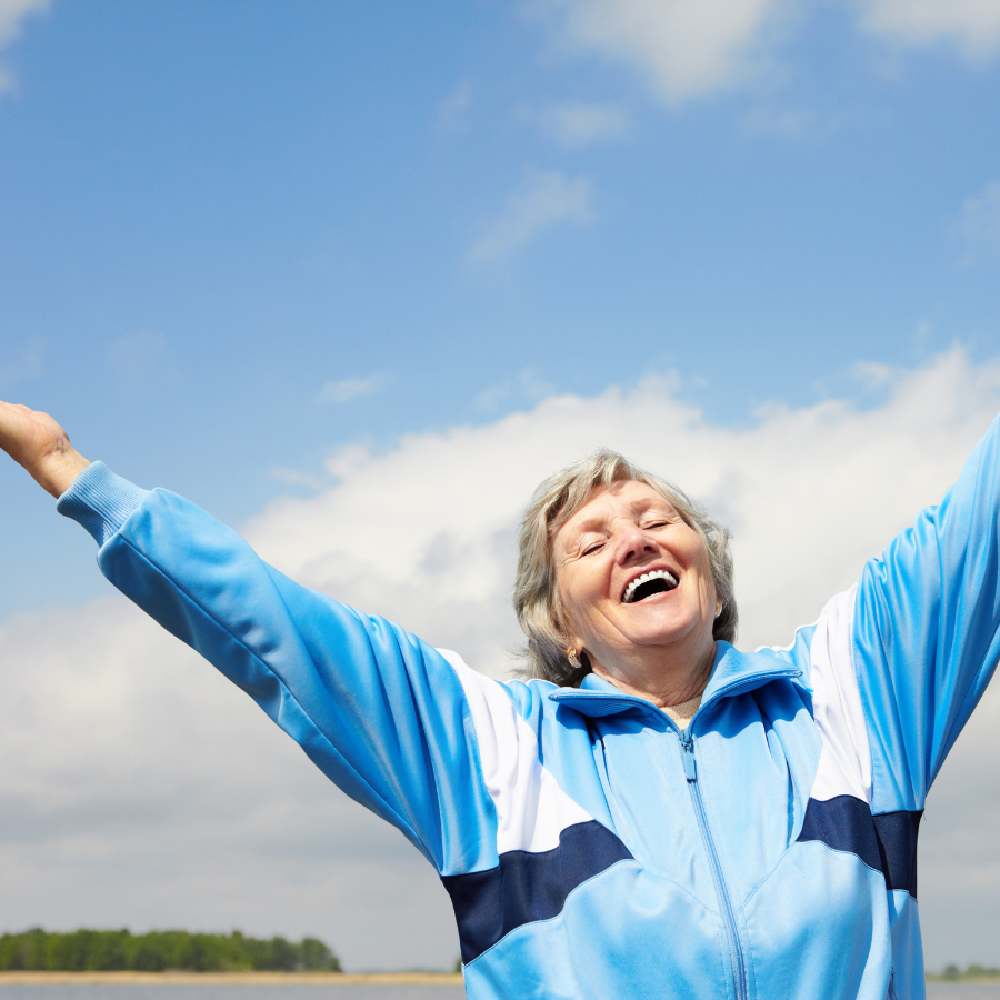 Smiling person in a light blue jacket with arms raised against a blue sky outdoors