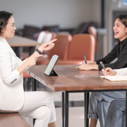 Two people talking across a table in a bright meeting room, one pointing while the other takes notes.