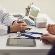 Doctor consulting with a patient at a desk, with a laptop, stethoscope, and tablet.