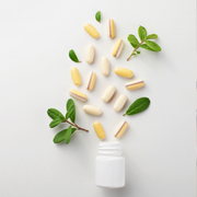 Assorted capsules and green leaves spilling from a white pill bottle on a white background