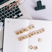 Scrabble tiles spelling “HEALTH INSURANCE” on an open planner beside a laptop and glasses