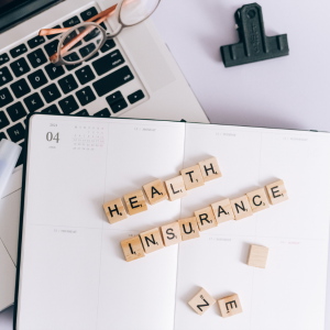 Scrabble tiles spelling “HEALTH INSURANCE” on an open planner beside a laptop and glasses