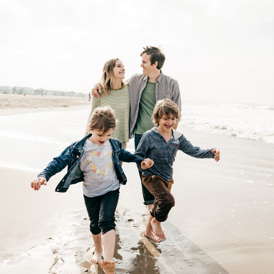 Two adults and two children walking barefoot on a beach, smiling and holding hands