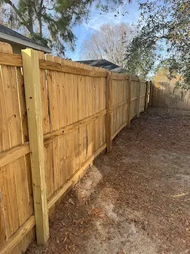 Wooden fence in a backyard, with a curved design, set on a bed of dry leaves, under a partly cloudy sky.
