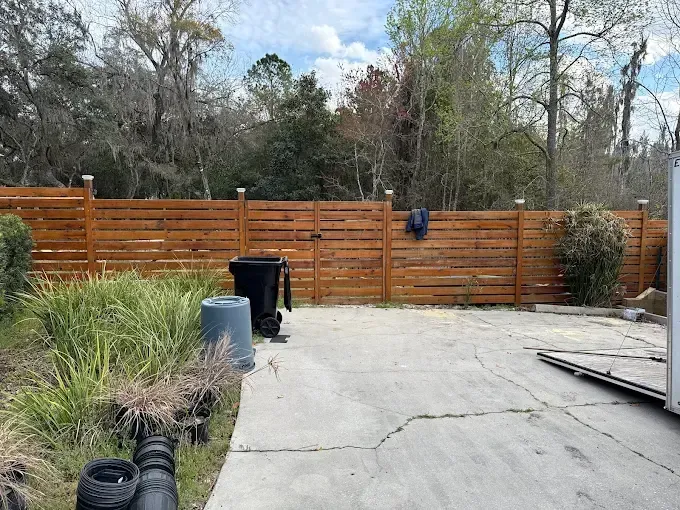 Backyard with stained wood fence, concrete patio, trash cans, and foliage. Trees in background, blue sky.